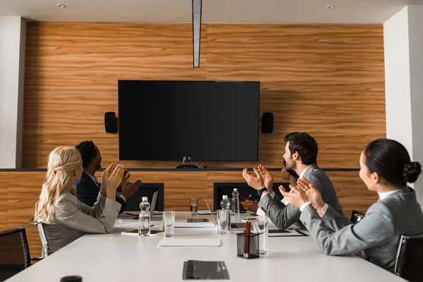 Junge multikulturelle Geschäftsleute applaudieren, während sie im Konferenzsaal sitzen und auf den LCD-Bildschirm an der Wand schauen — Stockfoto