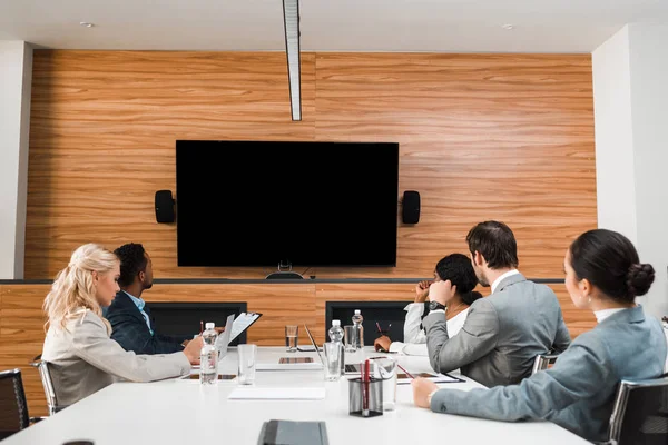 Young multicultural businesspeople sitting in conference hall and looking at lcd screen on wall — Stock Photo