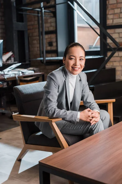 Feliz, atractivo asiático mujer de negocios sentado en sillón y sonriendo a la cámara - foto de stock