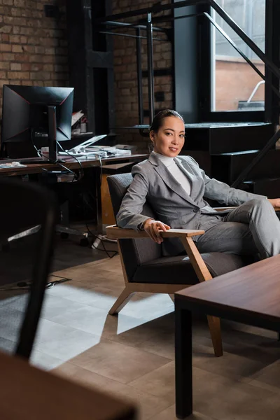 Hermosa asiático mujer de negocios sentado en sillón y sonriendo a cámara - foto de stock