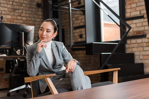 Привлекательная азиатская деловая женщина, сидящая в кресле и трогательная подбородок, глядя в сторону — стоковое фото