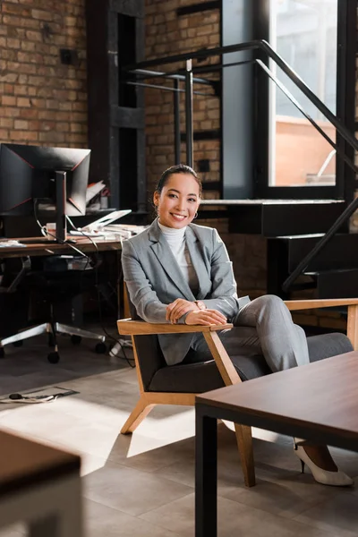 Избирательный фокус привлекательной азиатской предпринимательницы, сидящей в кресле и улыбающейся в камеру — стоковое фото