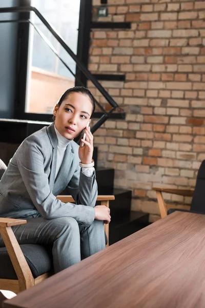 Réfléchie asiatique femme d'affaires assis dans un fauteuil, parler sur smartphone et détourner les yeux — Photo de stock