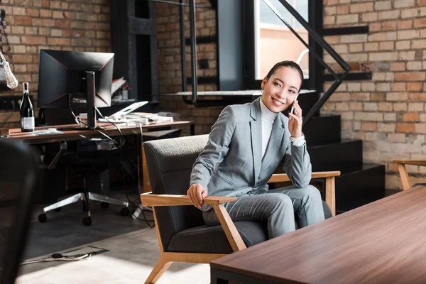 Alegre asiático mujer de negocios sentado en sillón y hablando en smartphone - foto de stock