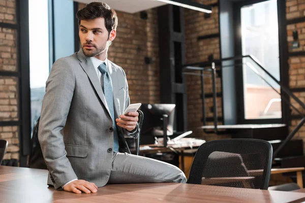 Hombre de negocios seguro y guapo sentado en el escritorio de la oficina, sosteniendo el teléfono inteligente y mirando hacia otro lado - foto de stock