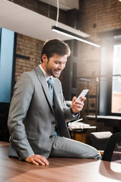 Fröhlicher, gutaussehender Geschäftsmann sitzt am Schreibtisch und plaudert auf dem Smartphone — Stockfoto