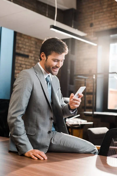 Alegre, hombre de negocios guapo sentado en el escritorio de la oficina y charlando en el teléfono inteligente - foto de stock