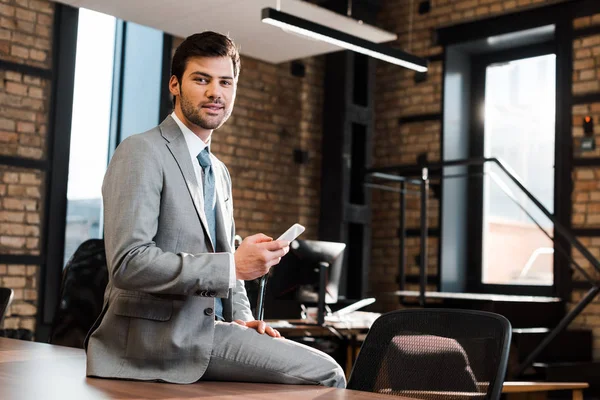 Bel homme d'affaires positif assis sur le bureau, tenant smartphone et regardant la caméra — Photo de stock