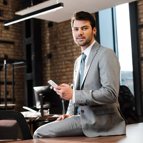 Bonito, homem de negócios sorridente sentado na mesa de escritório, segurando smartphone e olhando para a câmera — Fotografia de Stock