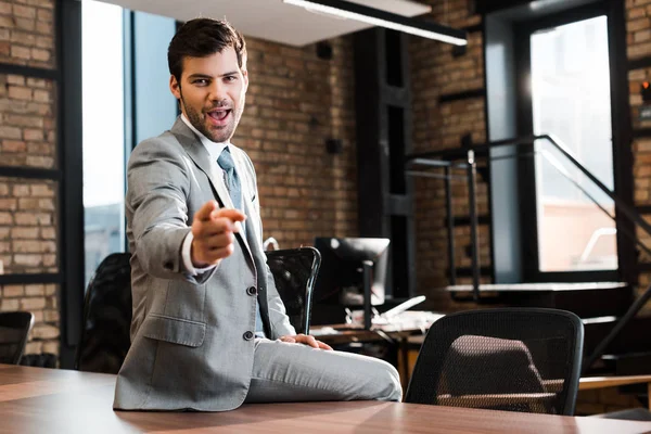Возбужденный, красивый бизнесмен сидит на рабочем столе и показывает пальцем в камеру — стоковое фото