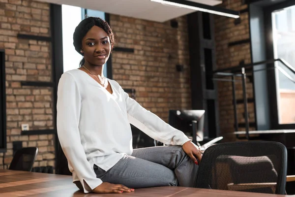 Alegre, mulher de negócios americana africana grávida sentada na mesa de escritório e sorrindo para a câmera — Fotografia de Stock