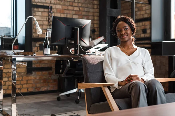 Feliz, embarazada mujer de negocios afroamericana sentada en sillón y sonriendo a la cámara - foto de stock