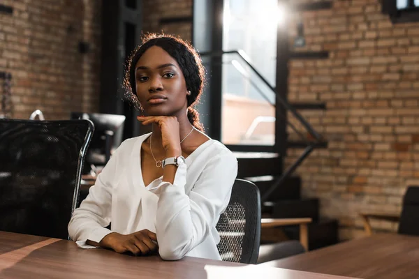 Atractiva, reflexiva mujer de negocios afroamericana sentada en el escritorio y mirando a la cámara - foto de stock