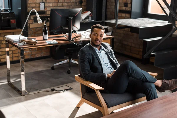 Sorridente empresário afro-americano sentado em poltrona e olhando para a câmera — Fotografia de Stock