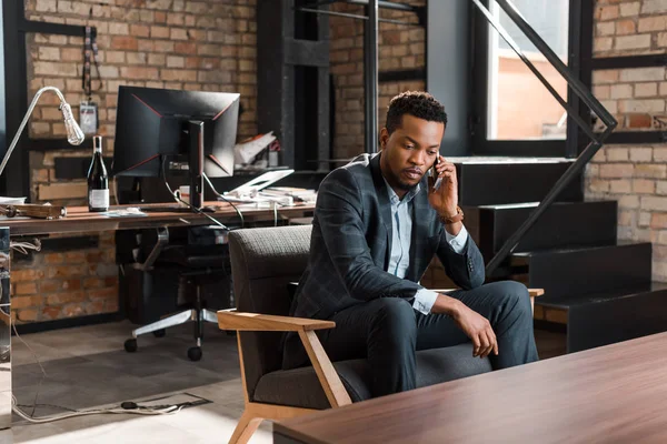 Серьезный африканский американский бизнесмен, сидящий в кресле и разговаривающий на смартфоне — стоковое фото