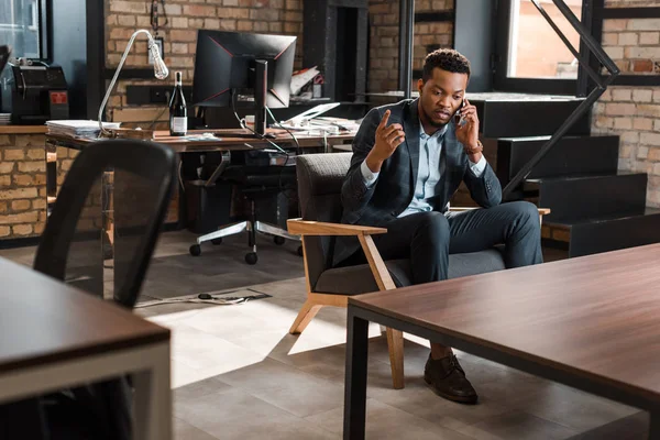 Hombre de negocios afroamericano serio sentado en sillón y mostrando gesto de idea mientras habla en el teléfono inteligente - foto de stock