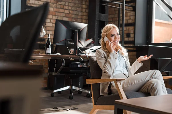 Selektiver Fokus einer lächelnden Geschäftsfrau, die mit offenem Arm im Sessel sitzt und mit dem Smartphone spricht — Stockfoto