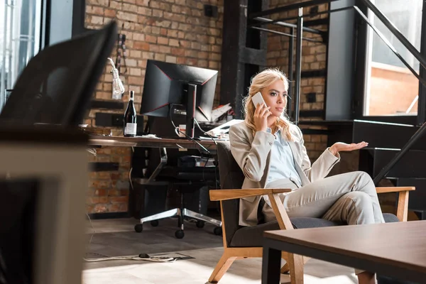 Selektiver Fokus einer seriösen Geschäftsfrau, die mit offenem Arm im Sessel sitzt und mit dem Smartphone spricht — Stockfoto