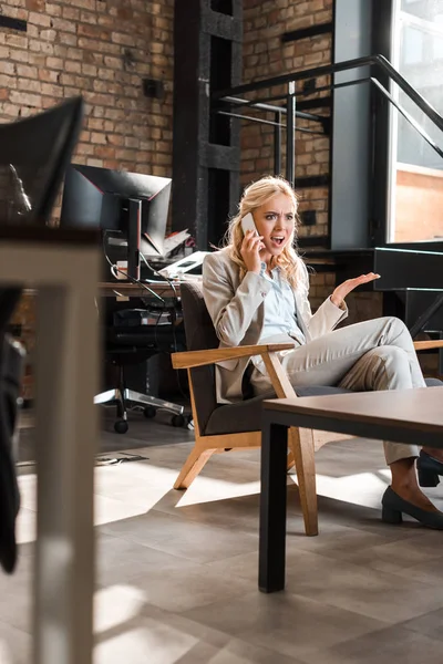 Selektiver Fokus einer wütenden Geschäftsfrau, die mit offenem Arm im Sessel sitzt und auf dem Smartphone schreit — Stockfoto