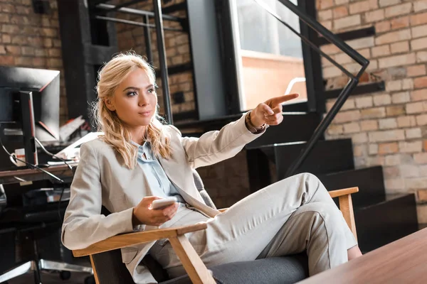 Atractiva mujer de negocios sonriente sentada en sillón, mirando hacia otro lado y señalando con el dedo - foto de stock