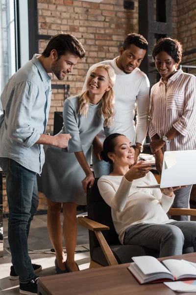 Cheerful multicultural businesspeople standing near happy asian colleague using laptop — Stock Photo