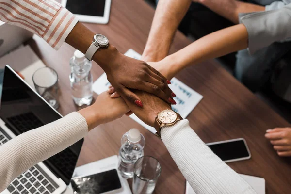 Visão parcial de empresários multiculturais segurando as mãos unidas acima da mesa com gadgets, vista superior — Fotografia de Stock