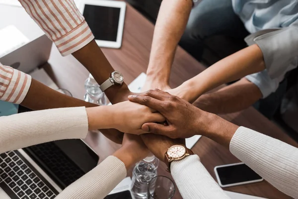 Cropped view of multicultural businesspeople holding joined hands above desk with gadgets, top view — Stock Photo