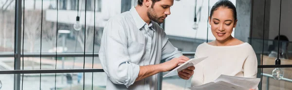 Plano panorámico de guapo hombre de negocios con digital tableta de pie cerca atractivo asiático colega celebración de papeles - foto de stock