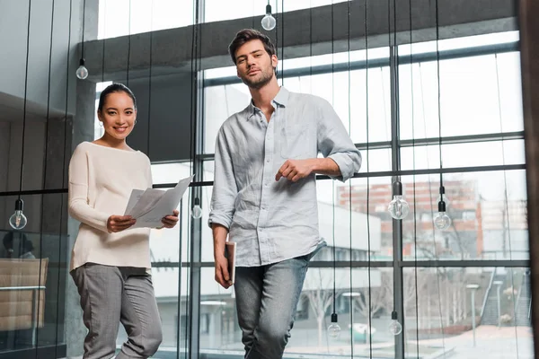 Pregnant asian businesswoman holding papers while standing near handsome colleague — Stock Photo