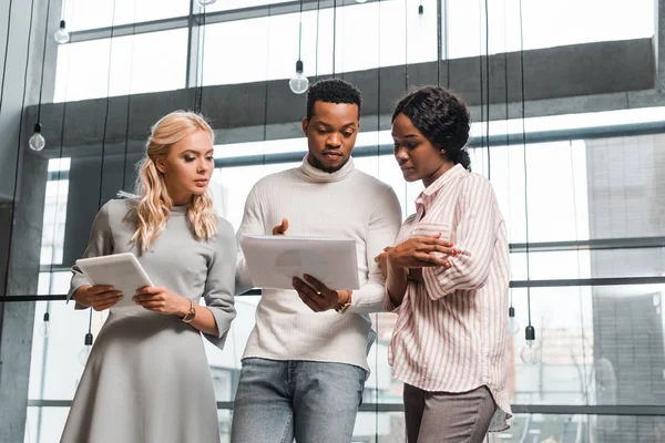 Young businesswoman holding digital tablet while standing near african american colleagues looking at documents — Stock Photo