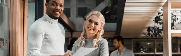 Plano panorámico de empresarios multiculturales felices mirando a la cámara mientras están de pie en la oficina - foto de stock