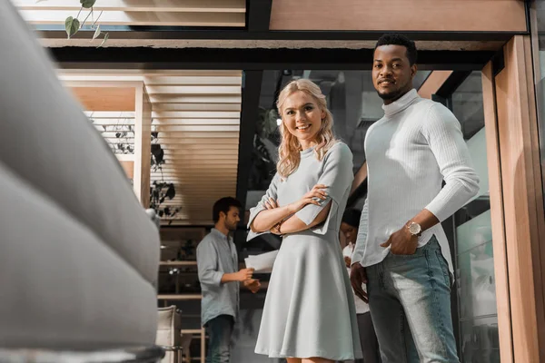 Selective focus of smiling multicultural businesspeople looking at camera while multicultural colleagues standing on background — Stock Photo