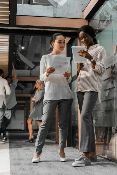 Concentrated african american and asian businesswomen with digital tablet and documents standing in office with multicultural colleagues on background — Stock Photo