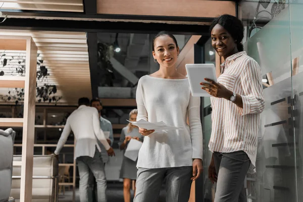 Lächelnde multikulturelle Geschäftsfrauen mit digitalem Tablet und Dokumenten, die in die Kamera schauen, während Kollegen im Hintergrund stehen — Stockfoto