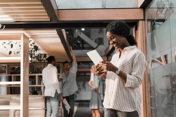 Mulher de negócios americana africana alegre usando tablet digital enquanto colegas multiculturais de pé em segundo plano — Fotografia de Stock