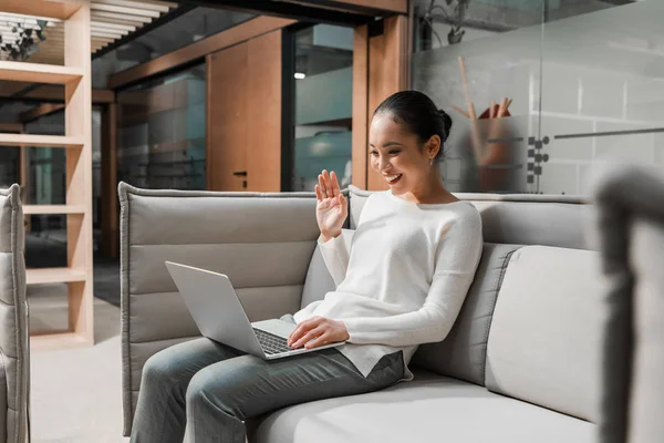 Щаслива азіатська бізнес-леді махає рукою, сидячи на дивані і маючи відеочат на ноутбуці — стокове фото