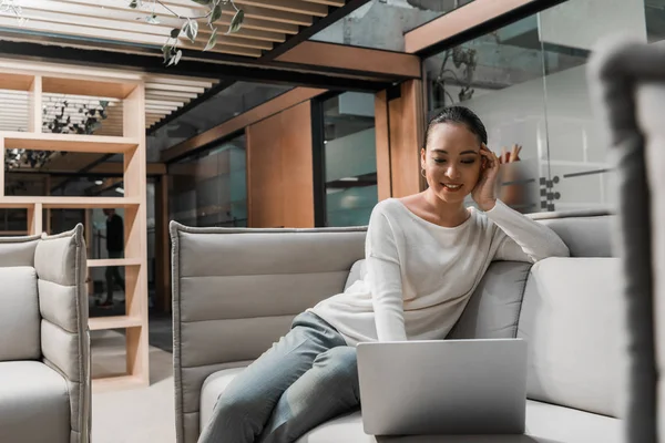 Selective focus of smiling asian businesswoman sitting on couch and using laptop — Stock Photo