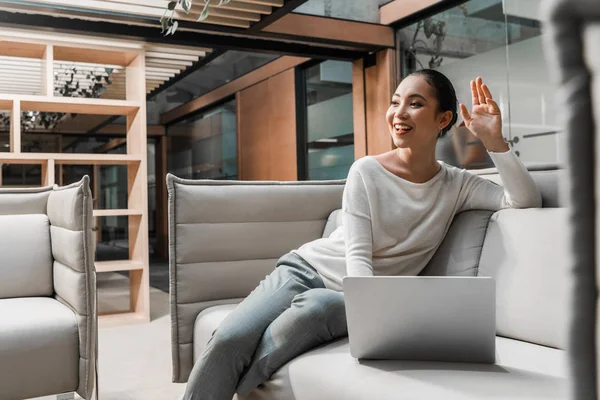 Вибірковий фокус усміхненої азіатської бізнес-леді махає рукою, сидячи на дивані біля ноутбука — стокове фото