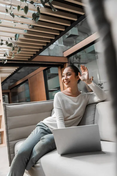 Enfoque selectivo de feliz asiático mujer de negocios saludando de la mano mientras está sentado en el sofá cerca de la computadora portátil - foto de stock