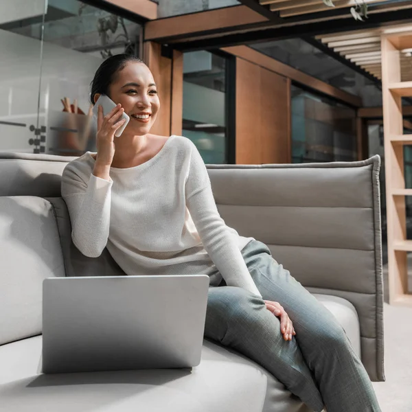 Happy asian businesswoman talking on smartphone while sitting on couch near laptop — Stock Photo