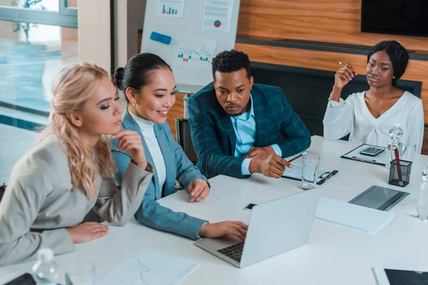 Serios empresarios multiculturales sentados en la sala de conferencias cerca de sonreír colega asiático utilizando el ordenador portátil - foto de stock