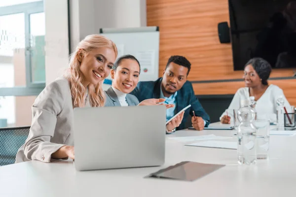 Des gens d'affaires multiculturels souriants assis dans la salle de conférence et regardant un ordinateur portable — Photo de stock