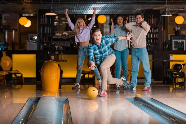 Joven guapo lanzando bola de bolos cerca de amigos multiculturales emocionados - foto de stock