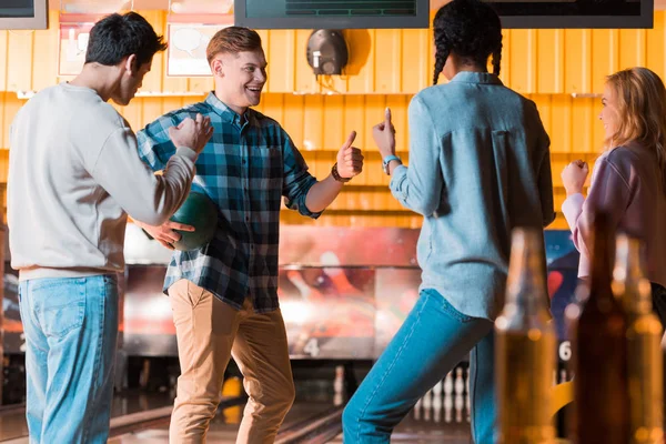 Selective focus of cheerful multicultural friends talking and showing thumbs up in bowling club — Stock Photo