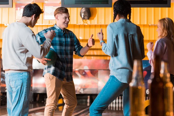 Selektiver Fokus fröhlicher multikultureller Freunde, die im Kegelclub miteinander reden und Daumen hoch zeigen — Stockfoto