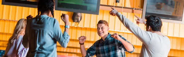 Панорамный снимок взволнованных мультикультурных друзей, улыбающихся и показывающих жест победителя в боулинг-клубе — стоковое фото