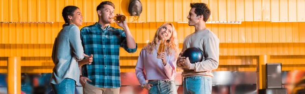 Panoramic shot of happy multicultural friend talking and clinking bottles of beer in bowing club — Stock Photo