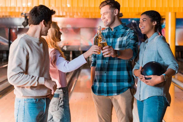 Happy multicultural friend talking and clinking bottles of beer in bowing club — Stock Photo