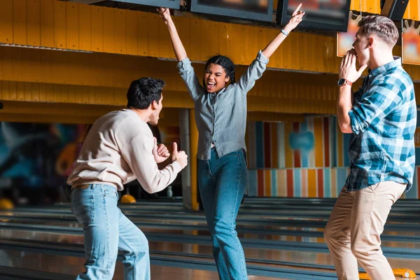 Happy african american girl standing with hands in air near multicultural friends showing thumbs up and screaming in bowling club — Stock Photo