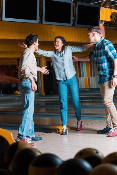 Feliz africano americano chica de pie con los brazos abiertos cerca alegre multicultural amigos en bowling club - foto de stock